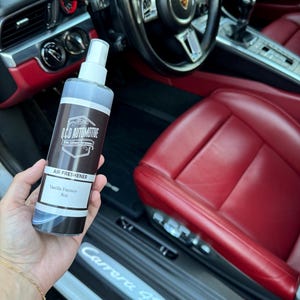 May include: A clear spray bottle with a black label that says "Q.C.D AUTOMOTIVE" and "AIR FRESHENER" with the scent "Vanilla Fantasy" 8oz. The bottle is being held in a hand in the interior of a white Porsche Carrera S.