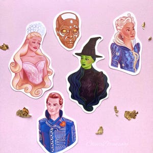 May include: A collection of die-cut stickers featuring characters from a musical. The stickers include a woman in a pink dress, a bronze-skinned face, a green-skinned witch, a woman in a blue coat, and a man in a blue jacket. Gold leaf accents the pink background.
