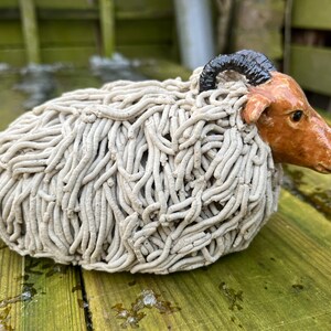 Könnte beinhalten: Keramik-Schafskulptur mit einem strukturierten, cremefarbenen, wolligen Körper. Das Schaf hat ein braunes Gesicht, schwarze Hörner und ruht auf einer Holzoberfläche. Die Skulptur ist ein dekoratives Objekt.