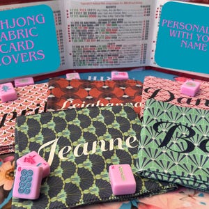 May include: Fabric card covers for Mahjong, personalized with names. The covers feature various patterns in red, green, and black, with names like "April", "Jeanne", and "Beth" displayed prominently. Pink Mahjong tiles are also present.