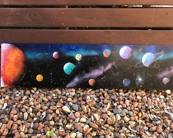 May include: A black canvas painting depicting a starry night sky with a galaxy of planets in various colors, including orange, pink, blue, and yellow. The planets are scattered across the canvas, creating a sense of depth and wonder.