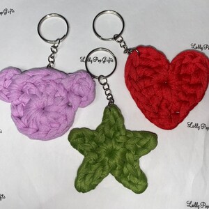 May include: Three crocheted keychains: a purple bear, a red heart, and a green star. Each keychain is attached to a silver key ring.