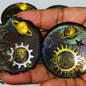 May include: Two round resin coasters with a space theme. Each coaster features a galaxy background with gold glitter, a planet with rings, stars, and a gear design.
