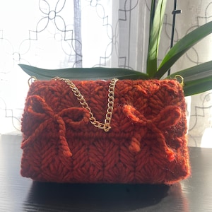 May include: A small, rust-colored knitted handbag with a gold chain strap and two large bows on the front.