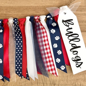 May include: A collection of colorful ribbons in red, white, and blue, with patterns including polka dots, paw prints, and gingham. A white tag with the word "Bulldogs" in black script is attached to the ribbons.