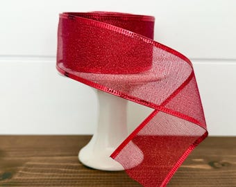 May include: A roll of red ribbon with a metallic sheen, displayed on a white pedestal. The ribbon has a mesh-like texture and a wired edge, perfect for crafting or gift wrapping. The ribbon is on a dark brown wooden surface.