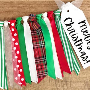 May include: A festive Christmas ribbon garland with red, green, white, and plaid ribbons. A white tag with the words "merry Christmas!" is attached to the garland. The ribbons are tied together and are on a wooden surface.