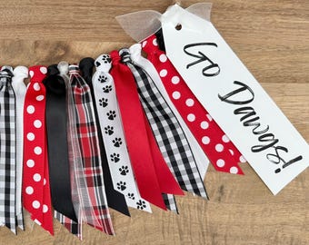 May include: A collection of colorful ribbons in red, black, and white patterns, including polka dots, gingham, and paw prints. A white tag with the words "Go Dawgs!" is attached to the ribbons.