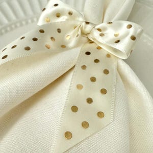 May include: A cream-colored napkin is adorned with a satin ribbon tied in a bow. The ribbon features gold polka dots, adding a touch of elegance. The napkin is folded and placed on a white plate, creating a decorative table setting.