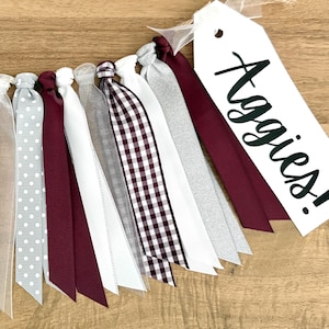 May include: A decorative banner with alternating maroon, white, and gray ribbons. Some ribbons have polka dots or a gingham pattern. A white tag with the word "Aggies!" in black script is attached to the banner.