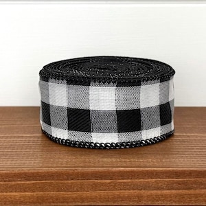 May include: A roll of black and white buffalo check ribbon. The ribbon is made of a woven fabric with a repeating pattern of large, square checks. The edges are finished with a black, decorative trim. The ribbon is sitting on a wooden surface.