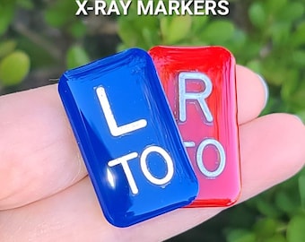 May include: Two small, rectangular x-ray markers. One is blue with a white "L" and "TO" printed on it. The other is red with a white "R" and "TO" printed on it. The markers are 1.25 inches tall and 0.75 inches wide.