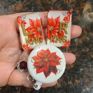 May include: A collection of Christmas-themed badge reels and name tags. The rectangular name tags feature red poinsettias, gold glitter lettering, and the letters "L" and "R". The round badge reel has a poinsettia design and the words "Merry Christmas".