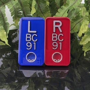 May include: A blue rectangular tag with the silver text L BC 91 and a circular cutout containing three small silver spheres, alongside a red rectangular tag with the silver text R BC 91 and a circular cutout containing three small silver spheres.