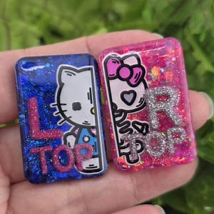 May include: Two rectangular resin charms with rounded corners. The left charm is blue with glitter and features a white cat character and the pink glitter text L TOP. The right charm is pink with glitter, a cat character, and silver glitter text R TOP.
