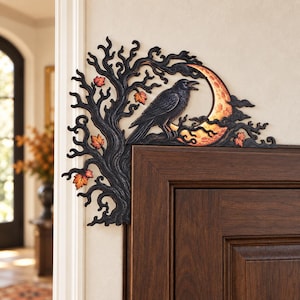 May include: A black decorative door corner accent featuring a gnarled tree silhouette with orange and red autumn leaves. A black raven is perched on a branch in front of a textured, glowing orange crescent moon, all set against a dark frame.