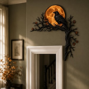 May include: A wall-mounted decorative piece featuring a black raven perched on a dark, gnarled tree branch. Behind the bird is a circular, glowing orange moon with a textured surface. Small, scattered orange leaves accent the dark, twisted branches.