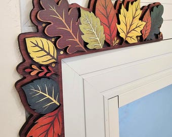 May include: A decorative wooden frame border featuring a layered cutout design of various autumn leaves in shades of deep red, golden yellow, sage green, and dark teal. The leaves are arranged in a dense, overlapping pattern along the edges.