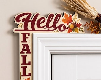 May include: An L-shaped wooden door corner sign featuring the dark red text Hello Fall. The design includes colorful autumn leaves and an acorn illustration. The sign is positioned on the top corner of a white door frame with a dried corn husk.