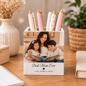 May include: A square white desk organizer featuring a printed photograph of a woman reading a book with two children. The text "Best Mom Ever" is printed below the photo, followed by a small heart and "LOVE, EMMA & NOAH".