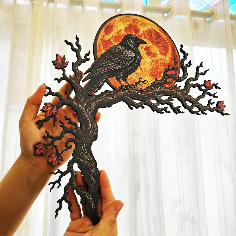 May include: A decorative wall hanging featuring a black raven perched on a dark, gnarled tree branch. Behind the bird is a large, circular orange moon with a textured surface. Small, reddish-orange autumn leaves are attached to the branch.