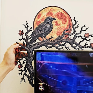 May include: A decorative wall accent featuring a black raven perched on a gnarled, leafless tree branch with small red leaves. Behind the bird is a large, circular, textured orange moon. The piece is designed to frame the corner of a flat screen.