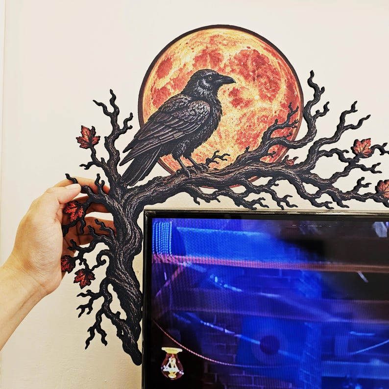 May include: A decorative wall accent featuring a black raven perched on a gnarled, leafless tree branch with small red leaves. Behind the bird is a large, circular, textured orange moon. The piece is designed to frame the corner of a flat screen.