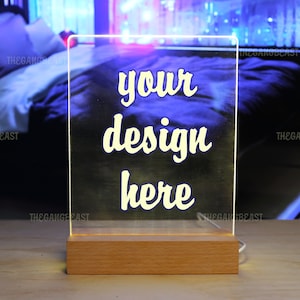 Op de afbeelding: Een rechthoekig, transparant acrylbord met een verlichte rand, voorzien van de witte tekst "your design here" in een cursief lettertype. Het acrylpaneel is gemonteerd op een rechthoekige, lichtgekleurde houten voet met een stroomkabel.