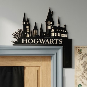 May include: A black wall-mounted door corner sign featuring a silhouette of the Hogwarts castle with illuminated windows. The word HOGWARTS is printed in white capital letters across the base of the castle structure, which is positioned on the top right corner of a door frame.
