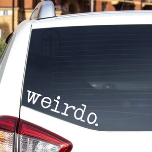 May include: A white vinyl decal applied to the rear window of a white vehicle. The decal features the word "weirdo." written in a clean, lowercase, sans-serif font. The text is positioned horizontally across the lower section of the glass.