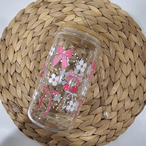 May include: A clear glass tumbler with a bamboo lid and straw. The tumbler is decorated with pink bows, white flowers, and red cherries. The tumbler sits on a woven, circular placemat.