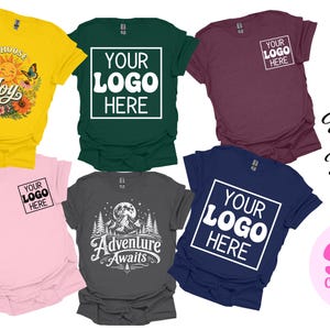 Peut inclure: Six t-shirts à manches courtes en jaune, vert foncé, bordeaux, rose clair, gris anthracite et bleu marine. Les motifs incluent 'Choose Joy' avec soleil et fleurs, 'Adventure Awaits' avec montagnes, et 'YOUR LOGO HERE' dans des cadres blancs. Texte : 'Design It Your Way'.