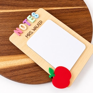 May include: A wooden memo board with a white writing surface, the word "NOTES" in colourful letters, and the name "MRS. WALKER" printed below. A red apple with a green leaf is attached to the bottom. The board is set on a wooden surface.