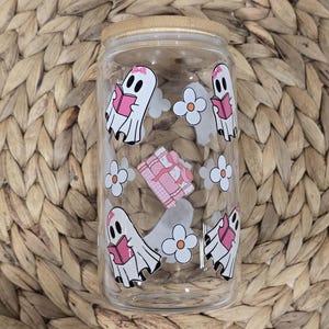 May include: A clear glass tumbler with a bamboo lid, decorated with white ghost illustrations reading pink books, white flowers, and a pink and white checkered design. The tumbler is set against a woven background.