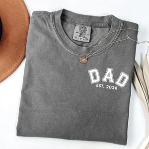 Peut inclure: Un t-shirt gris anthracite à manches courtes avec le texte blanc DAD et EST. 2026 dans une police universitaire sur la poitrine gauche. Le t-shirt a un col rond et une étiquette intérieure visible indiquant Comfort Colors, S-P-CH, 100% coton.