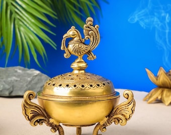 May include: A gold metal incense burner with a peacock design on the lid. The burner has three legs and is decorated with intricate details.