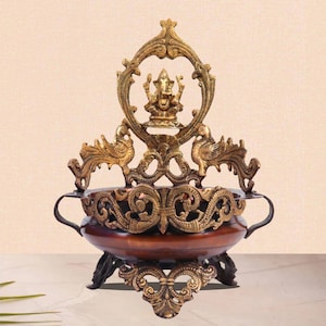 May include: A gold metal diya with intricate designs featuring peacocks and a Ganesha figure. The diya has a brown wooden bowl and a three-legged base.