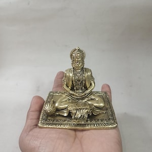 Puede incluir: Una estatua de metal dorado de una deidad sentada con una corona y joyas ornamentadas. La estatua está sentada en una plataforma decorativa con diseños intrincados.