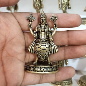 May include: A gold-colored metal figurine of a Hindu deity, Kurma Avatar, the second avatar of Vishnu, depicted as a turtle. The figurine is holding a chakra and a conch shell in its hands. The turtle shell is detailed with intricate carvings. The figurine is standing on a circular base with decorative designs.