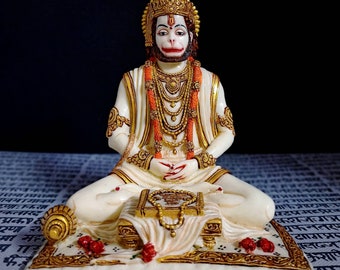 Może przedstawiać: Biała figurka hinduskiego bóstwa Hanuman siedzącego w medytacyjnej pozie. Ma na sobie złotą koronę, białą szatę i czerwoną i złotą naszyjnik. Trzyma książkę na kolanach i różaniec w dłoniach. Figurka stoi na białej podstawie ze złotą ozdobą.