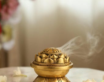 May include: A gold-colored incense burner shaped like a lotus flower with smoke rising from the top. The burner is sitting on a white marble surface.