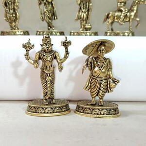 May include: Two gold-colored metal figurines on stands. The figurine on the left is a four-armed deity with a crown and a fierce expression. The figurine on the right is a man holding an umbrella and a round object.