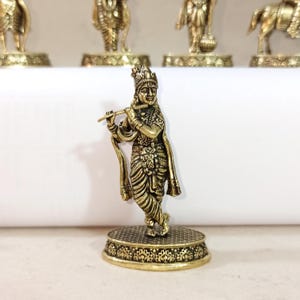 May include: A gold-colored metal figurine of a Hindu deity playing a flute. The figurine is standing on a decorative base with intricate details.