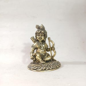 May include: A gold-coloured metal statue of a young boy sitting with a bow and arrow. The statue is on a base with a leaf design.