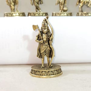 May include: A gold-colored metal figurine of a Hindu deity holding an axe and bow. The figurine is standing on a decorative base.