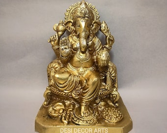 May include: A gold-colored brass statue of Ganesha, the Hindu god of wisdom and prosperity, sitting on a throne. The statue is intricately detailed and features a traditional design. The base of the statue is inscribed with the words "DESI DECOR ARTS".