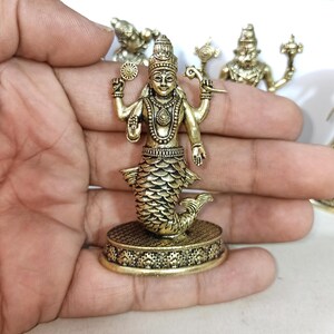 May include: A gold-colored metal figurine of a Hindu deity with a fish tail. The deity has four arms and is holding a conch shell, a discus, a lotus flower, and a mace. The figurine is standing on a circular base with intricate designs.