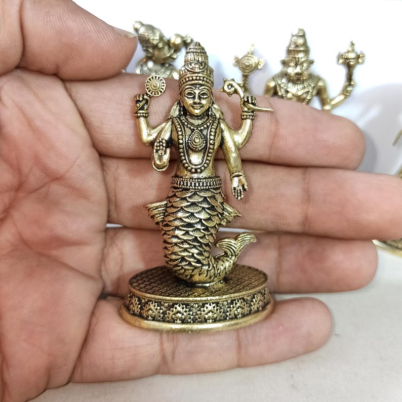 May include: A gold-colored metal figurine of a Hindu deity with a fish tail. The deity has four arms and is holding a conch shell, a discus, a lotus flower, and a mace. The figurine is standing on a circular base with intricate designs.
