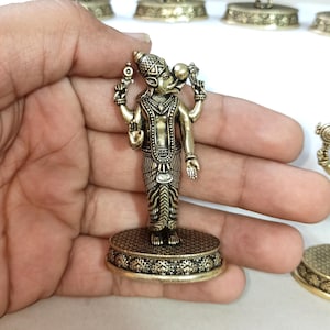 May include: A gold-colored metal figurine of a Hindu deity with four arms, holding various objects. The figurine is standing on a round base with intricate designs.
