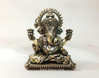 May include: A gold-colored metal statue of Ganesha, the Hindu god of beginnings, sitting on a throne with his trunk raised and holding a lotus flower in each hand. The statue is decorated with intricate details and has a halo behind the head. The statue is sitting on a white surface. "Desi Decor Arts" is written below the statue.